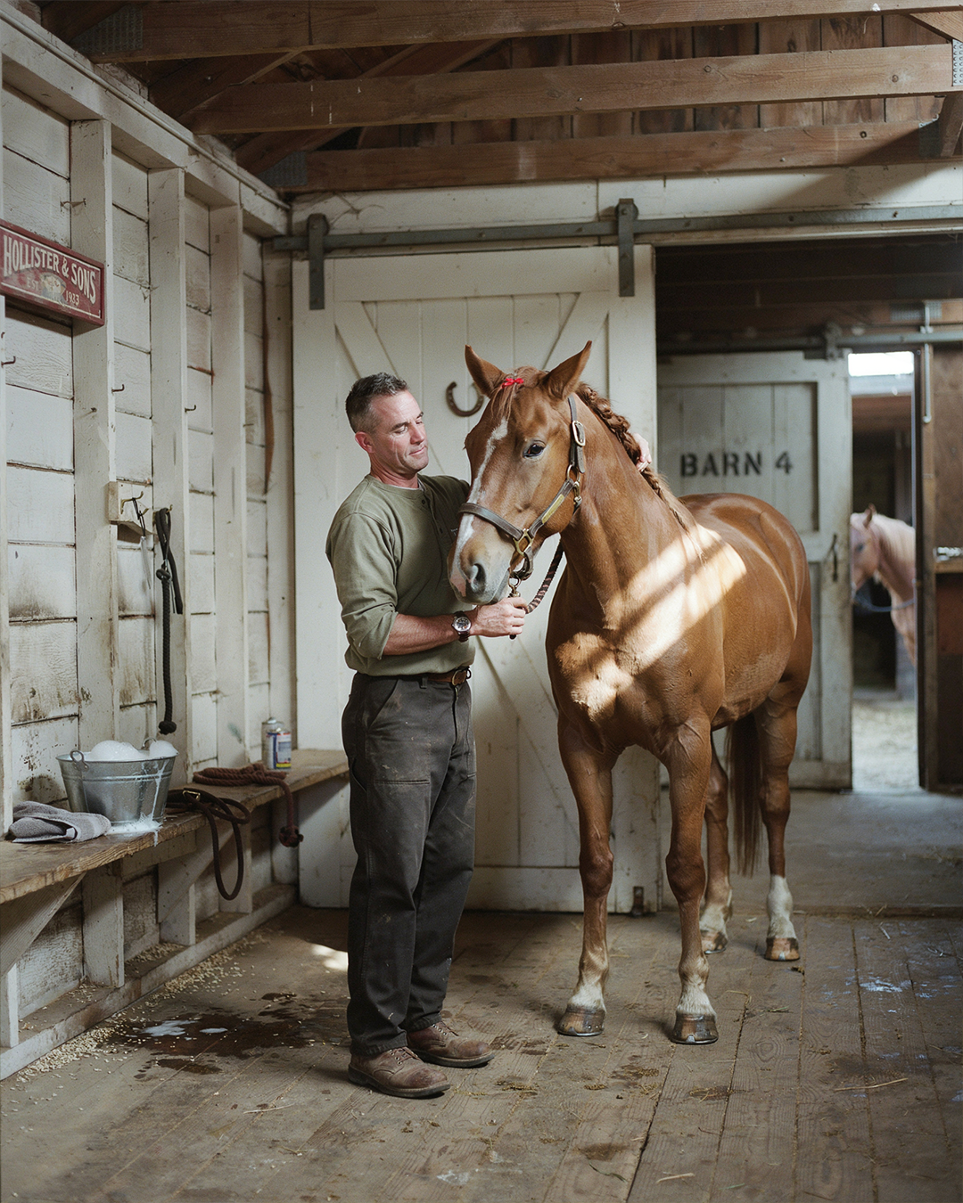 A military veteran quietly grooming a gentle therapy horse inside a beautifully restored American barn, warm morning sunlight streaming through wooden doors, peaceful atmosphere, authentic contemporary clothing, the horse calmly lowering its head toward the veteran, symbolizing trust, healing and hope through equine-assisted therapy.
