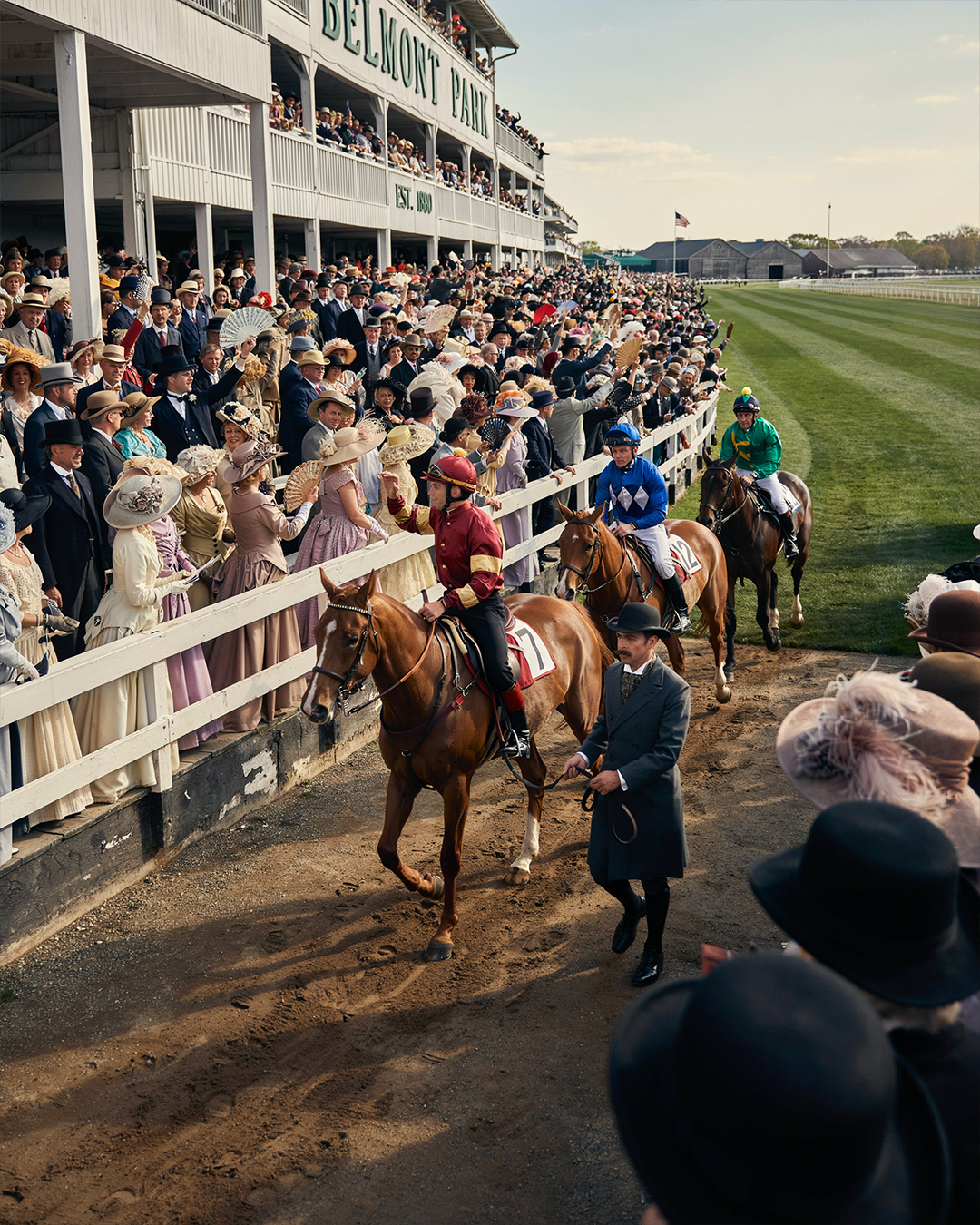Elegant Thoroughbred racehorses being paraded before a prestigious American racetrack around 1890, jockeys in authentic colorful silks, beautifully dressed spectators in Victorian fashion watching from the grandstand, warm afternoon sunlight.